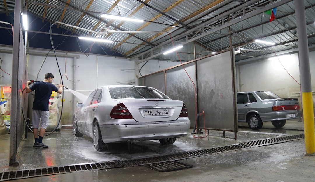 A man uses a pressure washer to clean a silver sedan in a covered car wash at night.