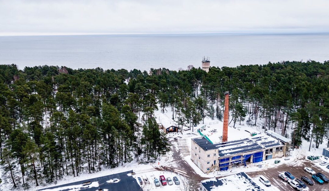 Aerial winter view of a snowy forest with an industrial area by the Baltic Sea in Ķesterciems, Latvia.