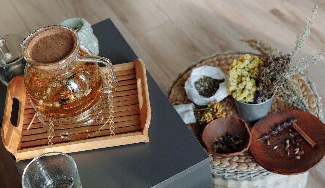 A glass teapot with herbal tea and dried flowers on a wooden tray indoors.