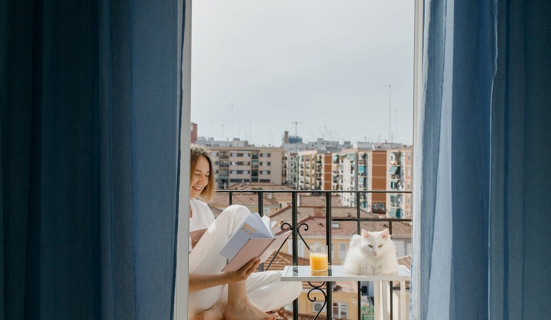 A woman enjoys reading on her balcony with a white cat and a glass of juice.