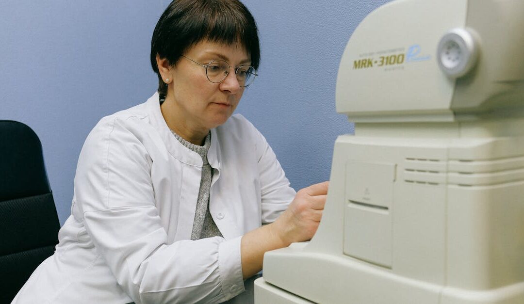 Serious middle aged eye doctor in eyeglasses working with automatic refkeratometer while sitting on chair leaned forward