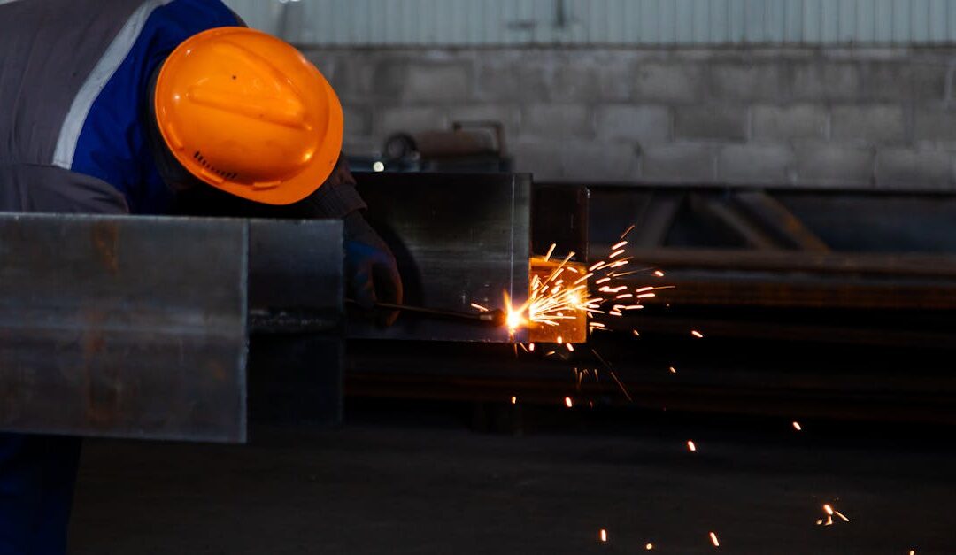 Worker wearing safety gear welds metal beam, emitting sparks in a factory setting.