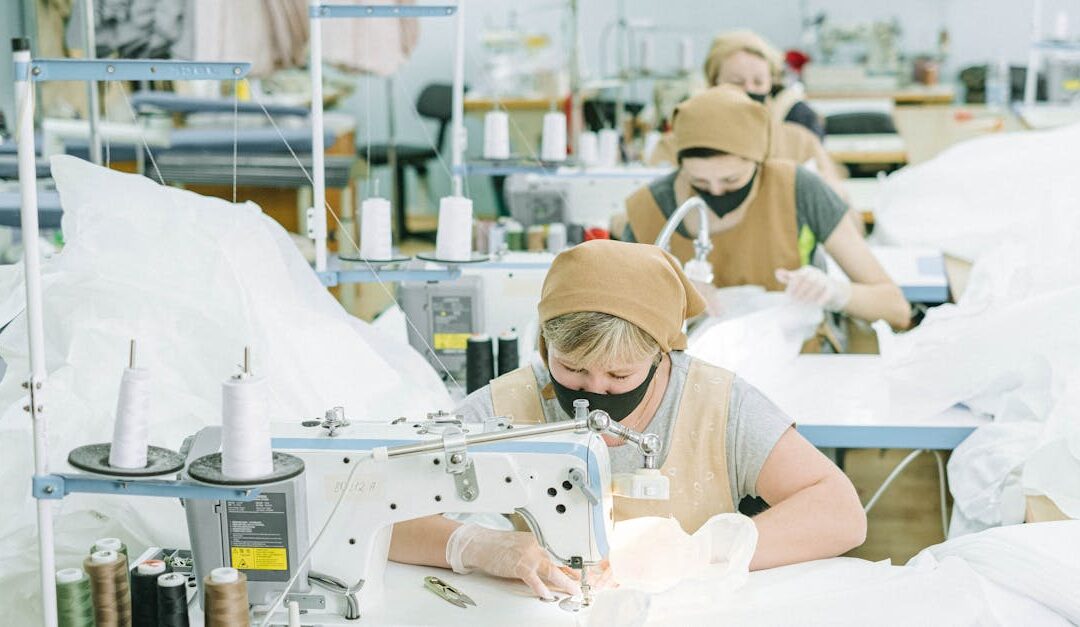 Women working on sewing machines in an industrial textile workshop, wearing protective masks and aprons.