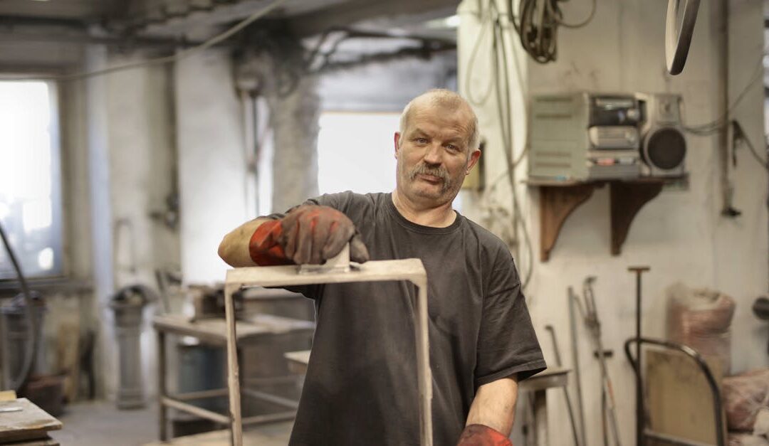 Pleased senior white hair craftsman in heavy duty gloves looking at camera while showing fabricated detaIl in workshop