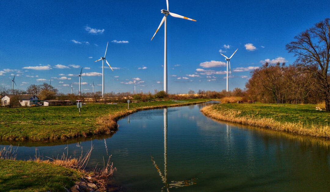 A serene landscape with wind turbines reflecting over a river under a clear blue sky.