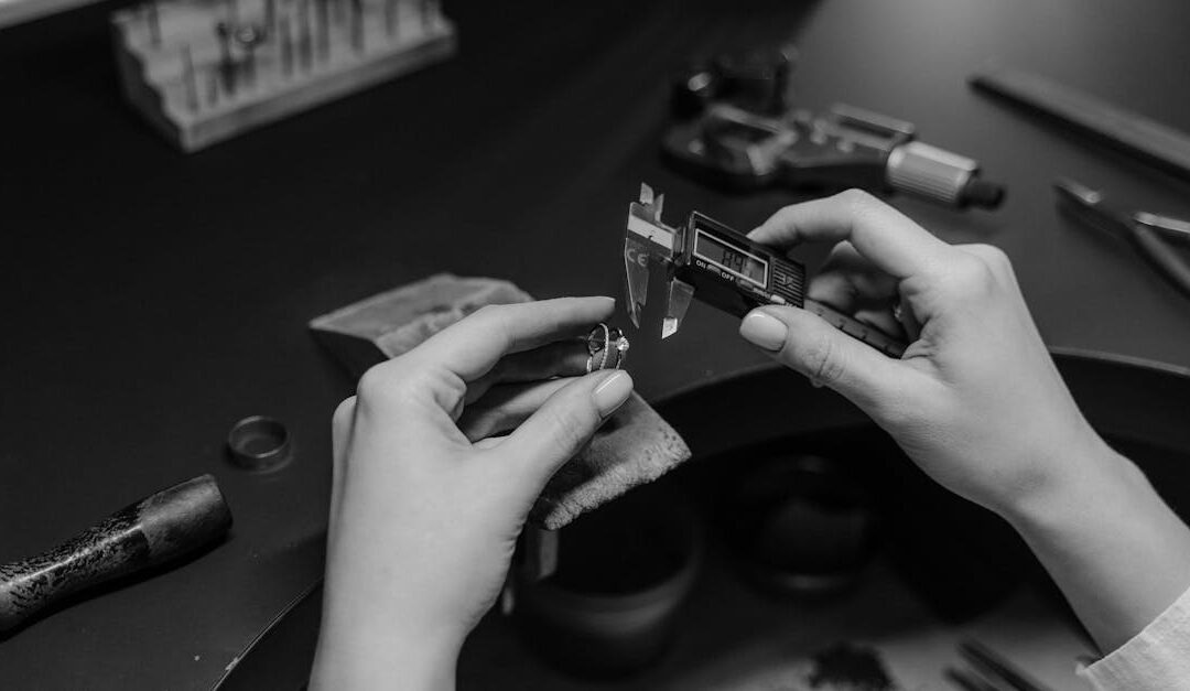 Black and white photo of a jeweler measuring a ring with a digital caliper in a workshop setting.